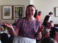 Eine sorbische Wirtin in Bautzen empfängt ihre Gäste, eine Reisegruppe aus Sachsen-Anhalt. Sie trägt sorbische Alltagstracht und breitet zum Willkommen die Arme aus.
